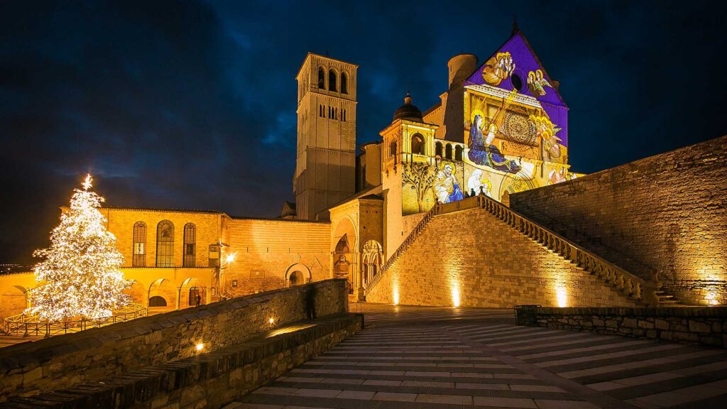 Basilica di San Francesco ad Assisi illuminata con proiezioni natalizie e albero di Natale acceso.